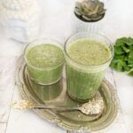 Two glasses of green protein-packed banana spinach smoothie on a silver display tray with a spoonful of oats, raw spinach on the side, and a plant in the background.