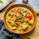Jackfruit Thai curry with onions and red pepper in a clay dish. A blue tea towel lying infrom of the dish and a bowl of basmati rice in the background.