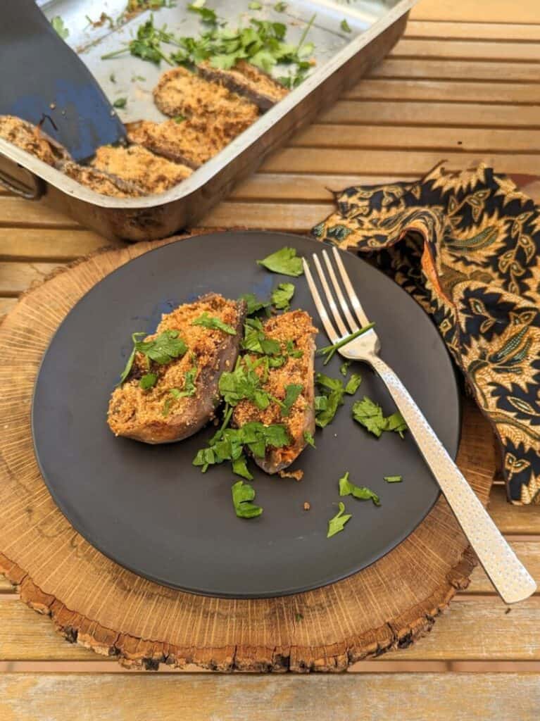 A plate with two sweet paprika Spanish-style stuffed eggplants, sprinkled with fresh parsley on top. To the right, there is a decorative black patterned cloth, and in the background, a tray half-full of more stuffed eggplants.