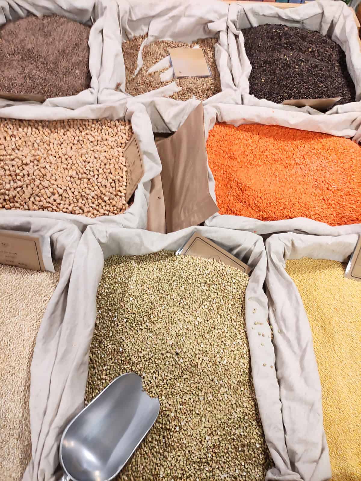 Bags of various dried pulses and grains, including red lentils, black beans, chickpeas, and seeds, displayed in a market with a metal scoop nearby.