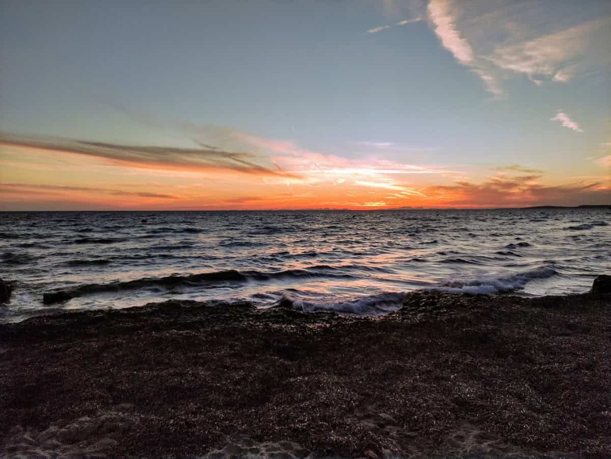A serene sunset over the ocean with soft waves, a dark shoreline, and vibrant orange and purple hues in the sky
