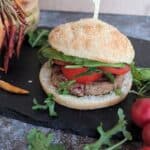 A plant-based bean burger with a jackfruit served in a whole-grain gluten free bun. The burger is topped with fresh avocado slices, a thick slice of tomato, and leafy greens, likely arugula, adding a fresh and vibrant look. The burger is placed on a black slate surface with some arugula leaves and radishes on the side, creating a rustic presentation. Three cherry tomoties are seen on the right and dry chili peppers on the left in the background.