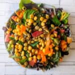 A vibrant salad featuring lupini beans, a bed of mixed green lettuce, including spinach. The salad also contains homemade pickled capers, diced tomatoes, pitted olives, carrot strips, cucumber slices, and avocado, creating a colorful and nutritious dish.