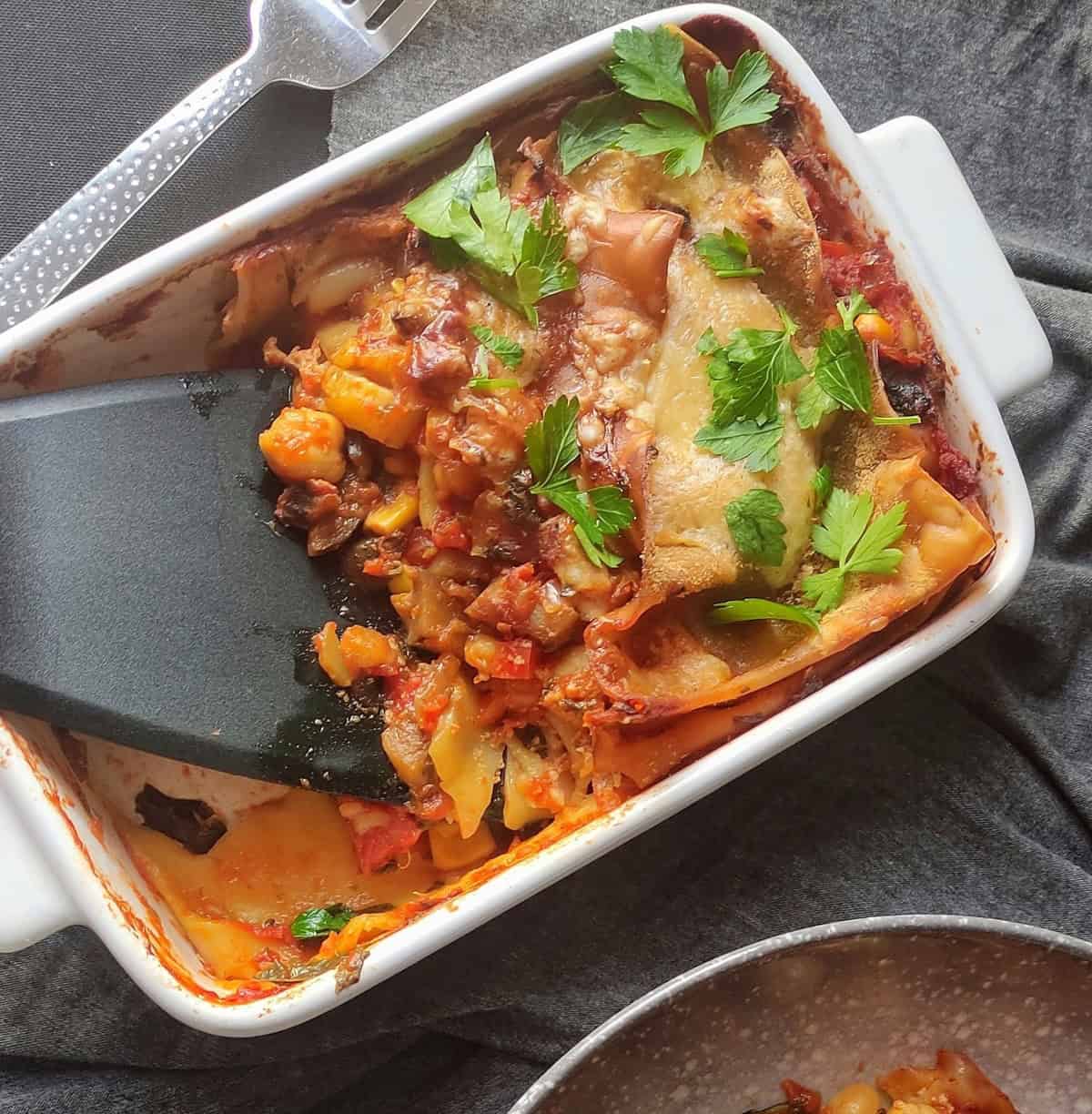 A white tray of chickpea and vegetable lasagna with a serving spatula dipped into it, dishing up a portion ready to serve.
