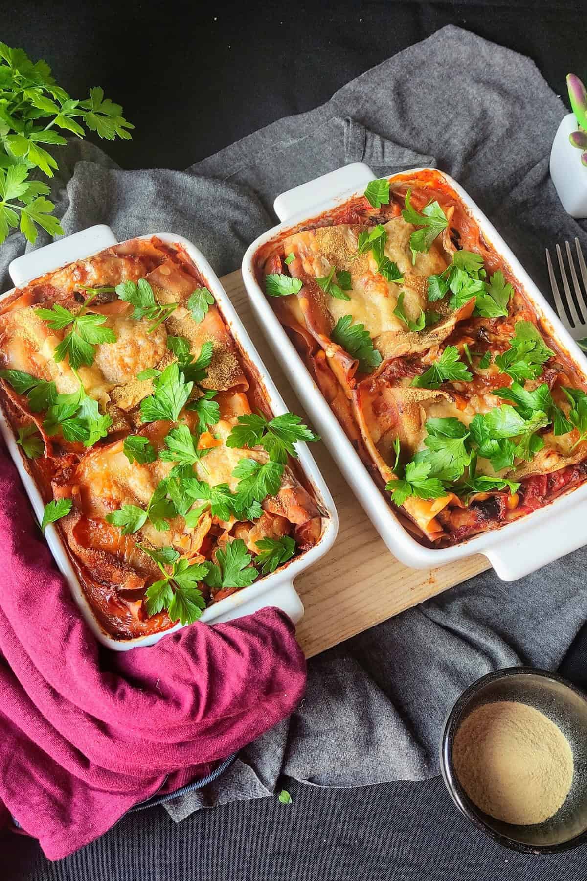 Two rectangular white baking dishes filled with chickpea and vegetable lasagna, topped with fresh parsley, placed on a dark gray cloth with a burgundy napkin nearby. A small bowl of nutritional yeast and a potted succulent are in the background for decoration.