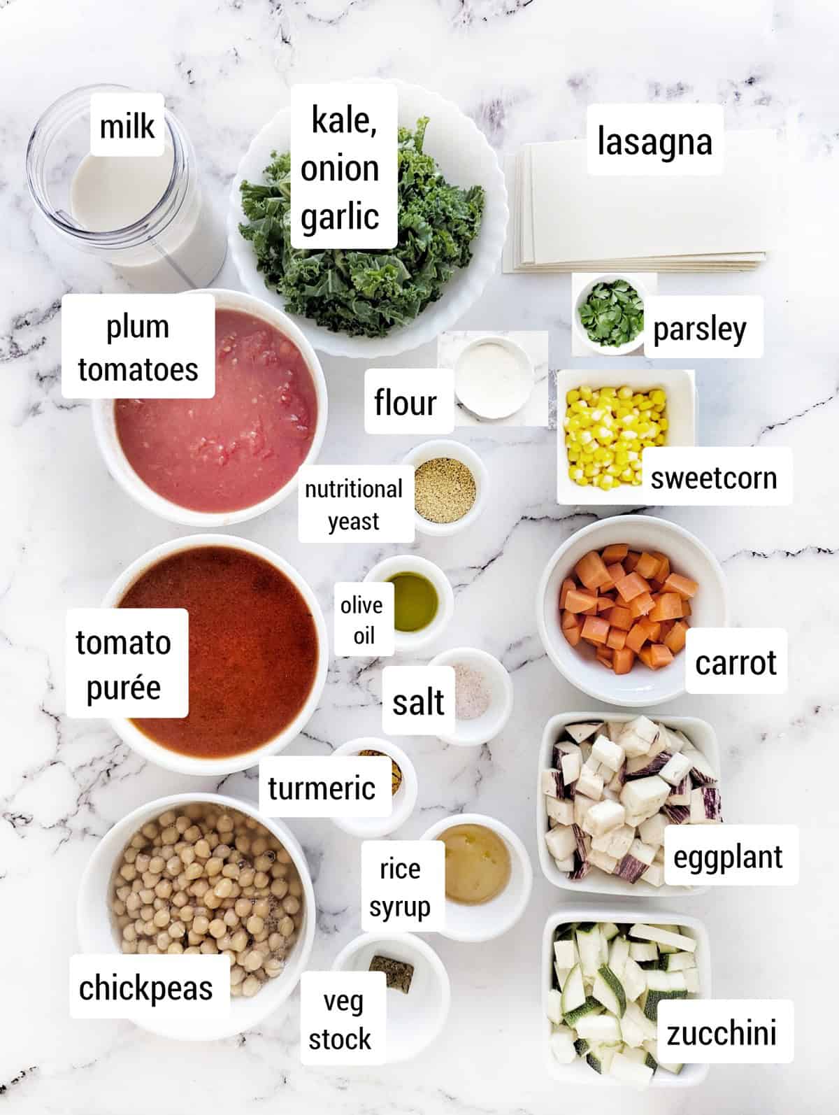 Overhead view of chickpea lasagna ingredients labeled on a white marble surface, including milk, kale, onion, garlic, lasagna sheets, parsley, sweetcorn, plum tomatoes, flour, nutritional yeast, tomato purée, olive oil, salt, turmeric, carrot, eggplant, zucchini, rice syrup, vegetable stock, and chickpeas.