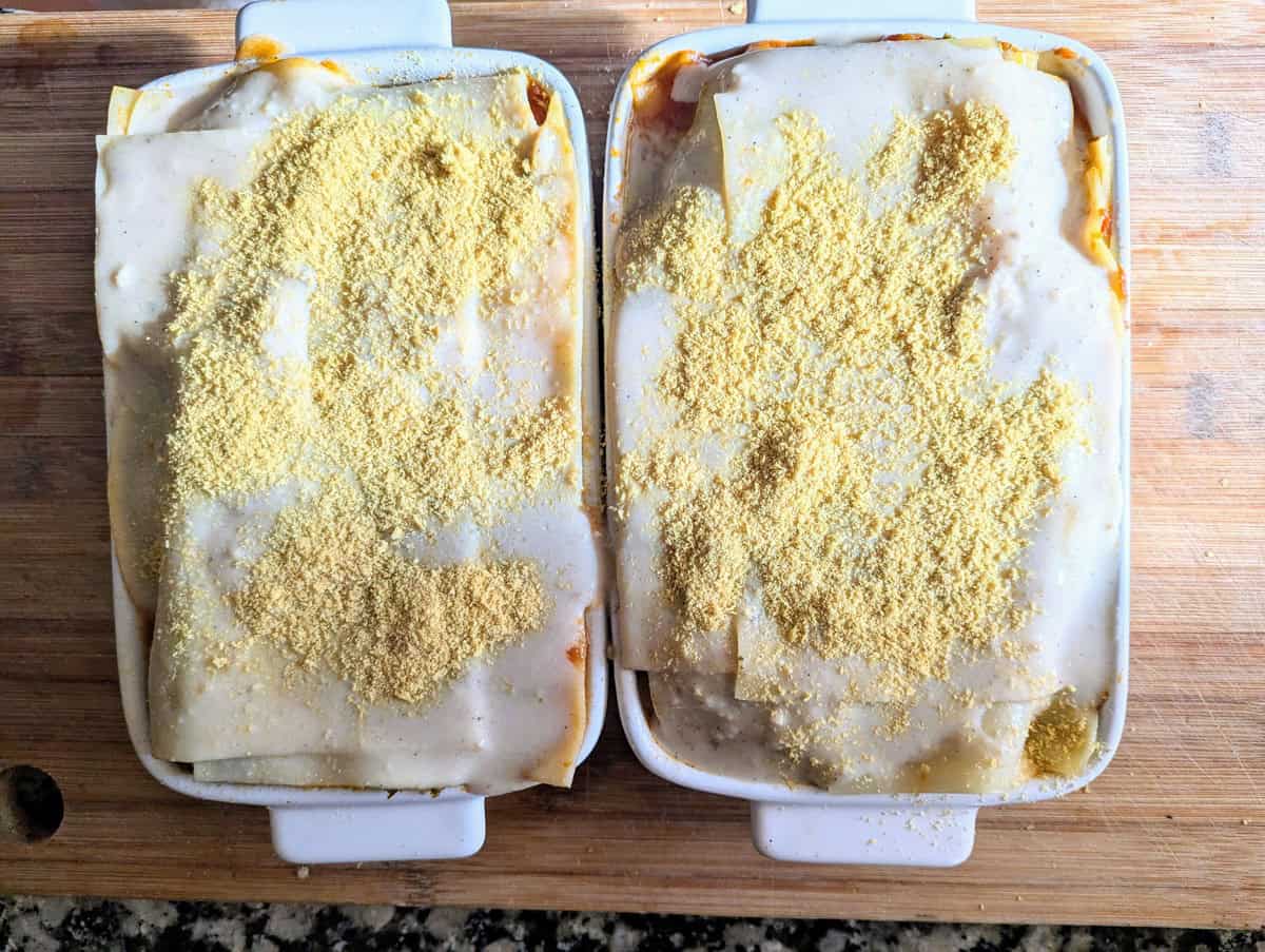 Two white trays of chickpea and vegetable lasagna placed on a brown board. The lasagna is topped with pasta sheets and nutritional yeast, ready to go in the oven.