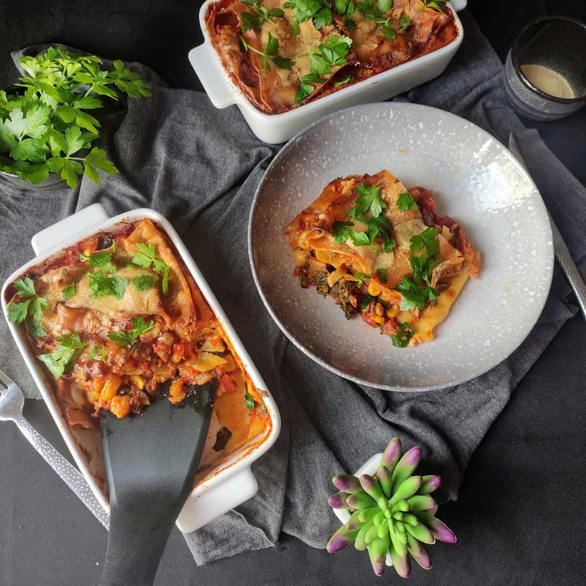 Two white trays of chickpea and vegetable lasagna are displayed, with a serving spatula dipped into one, dishing up a portion ready to serve. A plate with a single serving sits nearby, while another full white tray is visible in the background.