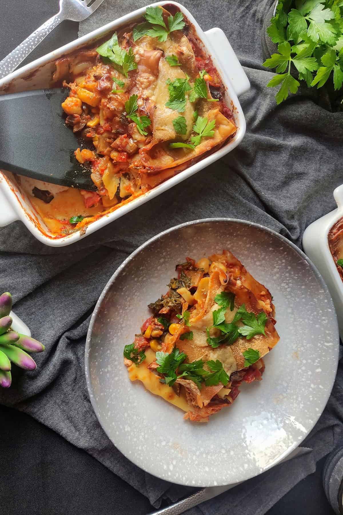 A white tray of chickpea and vegetable lasagna with a serving spatula dipped into one, dishing up a portion ready to serve. A plate with a single serving sits nearby.