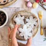 A close-up of a festive plate with three vegan mince pies topped with snowflake-shaped pastry lids dusted generously with powdered sugar. A hand holds one pie delicately. Surrounding the scene are a gold fork, a bowl of dried fruit, and Christmas ornaments on a marble surface, creating a cozy holiday atmosphere.