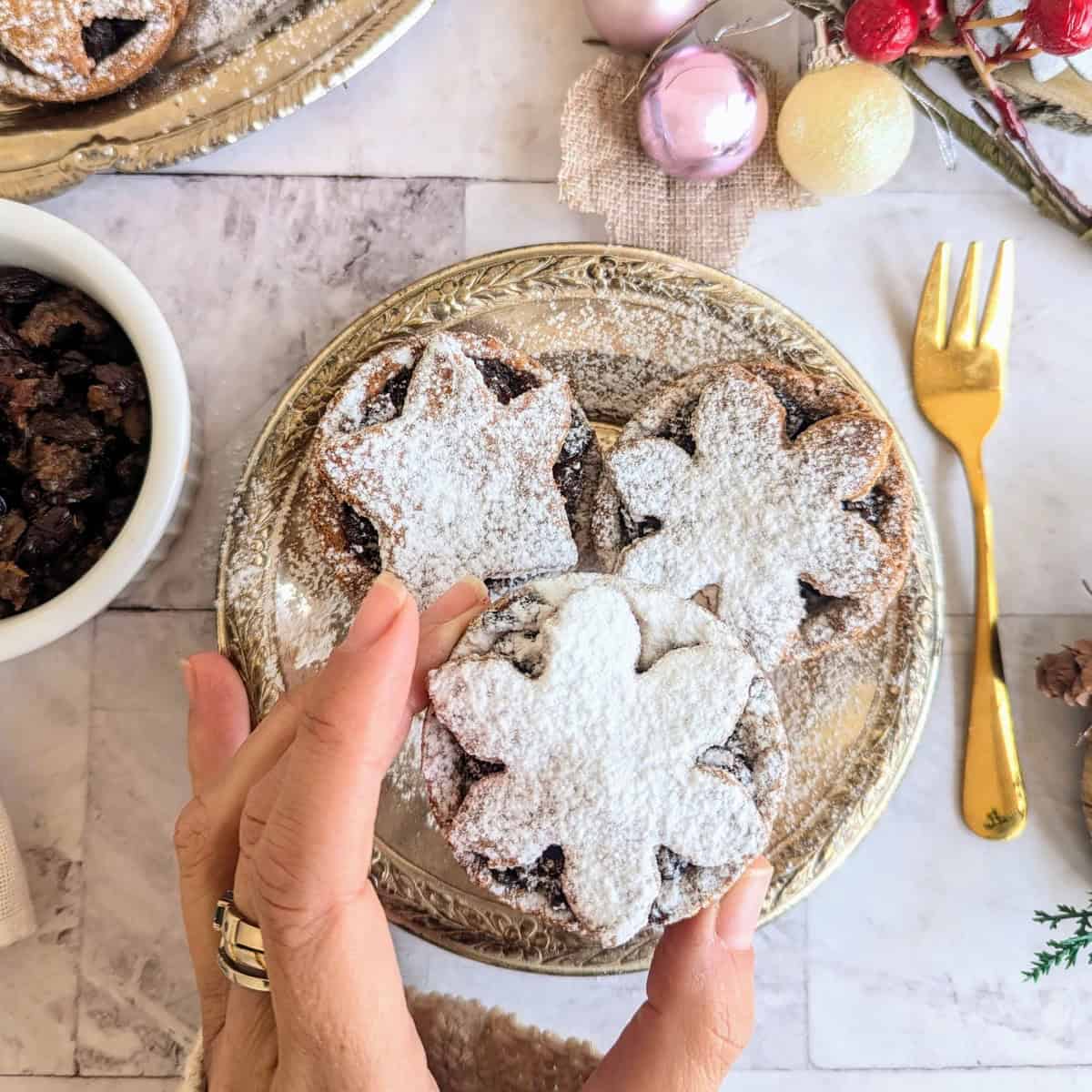 A close-up of a festive plate with three vegan mince pies topped with snowflake-shaped pastry lids dusted generously with powdered sugar. A hand holds one pie delicately. Surrounding the scene are a gold fork, a bowl of dried fruit, and Christmas ornaments on a marble surface, creating a cozy holiday atmosphere.