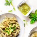 A dish of whole-wheat penne pasta coated in a vibrant green chili avocado pesto, garnished with fresh basil leaves. The pasta is served on a speckled grey ceramic plate, which rests on a round wooden board. A soft, off-white linen napkin drapes across the scene, adding a rustic touch. In the background, a glass bowl filled with additional pesto and a potted green plant enhance the natural, fresh aesthetic.