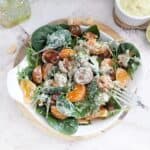 A fresh salad served in a white bowl, featuring black cherry tomatoes, chopped dates, spinach, arugula leaves, mandarin orange segments, and walnuts, all drizzled with a creamy lime, avocado, and cashew dressing. The bowl sits on a wooden board, accompanied by a silver fork. A small ramekin of extra dressing, a halved lime, and a glass bottle in the background. Cashew nuts are scattered on the wooden board.