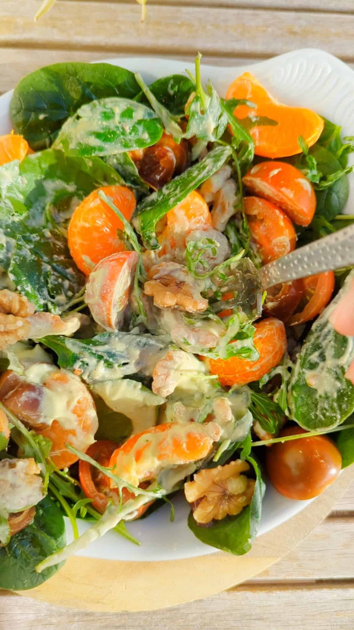 A close up view of a fresh salad served in a white bowl, featuring black cherry tomatoes, chopped dates, spinach, arugula leaves, mandarin orange segments, and walnuts, all drizzled with a creamy lime, avocado, and cashew dressing. A fork is digging into the salad.