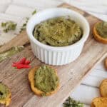 A creamy chili avocado pesto served in a white bowl on a rustic wooden board, accompanied by crispy golden crostini. The pesto has a rich green hue with flecks of spices, spread onto the bread slices. A knife with a generous dollop of pesto rests on the board, while a red chili pepper and fresh parsley adds to the scene.
