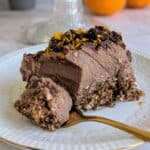 A slice of no-bake chocolate orange mousse cake on a white plate, with a creamy chocolate filling, crunchy nutty oat base, and topped with chocolate chunks and grated orand rind. The whole cake is displayed on a glass stand in the background, with fresh oranges.
