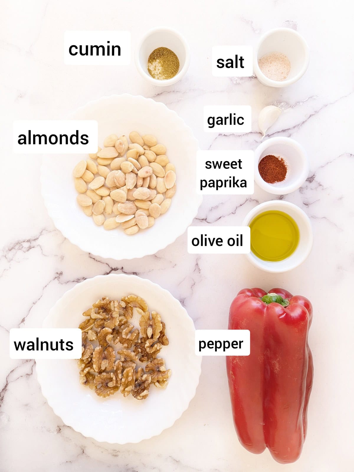Ingredients for muhammara dip arranged on a white marble surface, including a red pepper, walnuts, almonds, olive oil, garlic, cumin, sweet paprika, salt, and labeled bowls of spices.