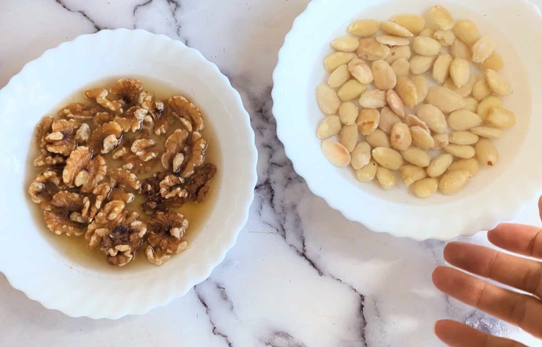 Walnuts in a white bow lin water and raw almonds in water in a white bowl.