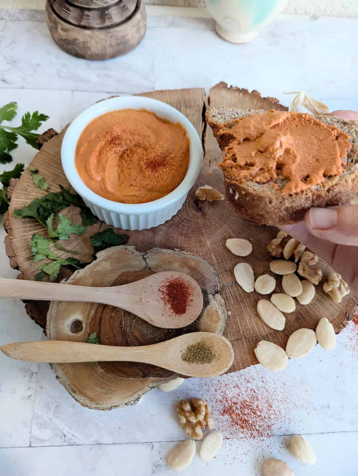 Muhammara dip in a white bowl, garnished with paprika, surrounded by walnuts, almonds, garlic, parsley, and wooden spoons filled with paprika and cumin, a hand holding bread ,all displayed on a rustic wooden board.
