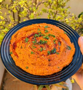 A middle eastern red pepper dip, muhammara sprinkled with parsley and sweet paprika. A plant is burred in the background.