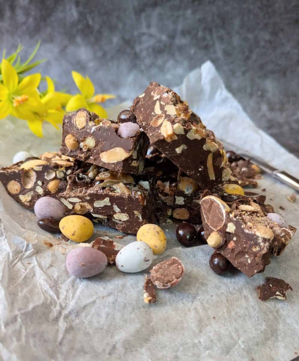 Close-up of stacked Rocky Road clusters with mini eggs and nuts in front, yellow flowers behind, and a faded knife on the right.