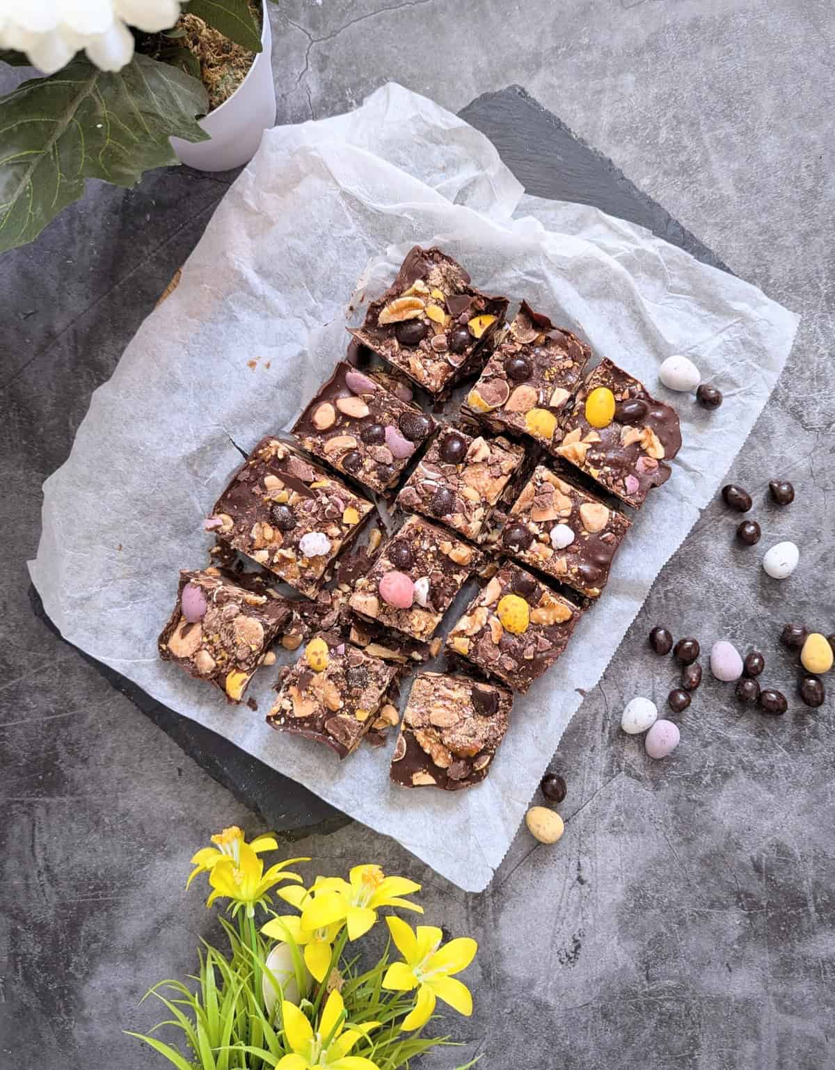 Rocky Road Easter chocolate clusters sliced into squares on parchment paper, topped with mini eggs, chocolate coated peanuts, and nuts surrounded by spring flowers and Easter candy.