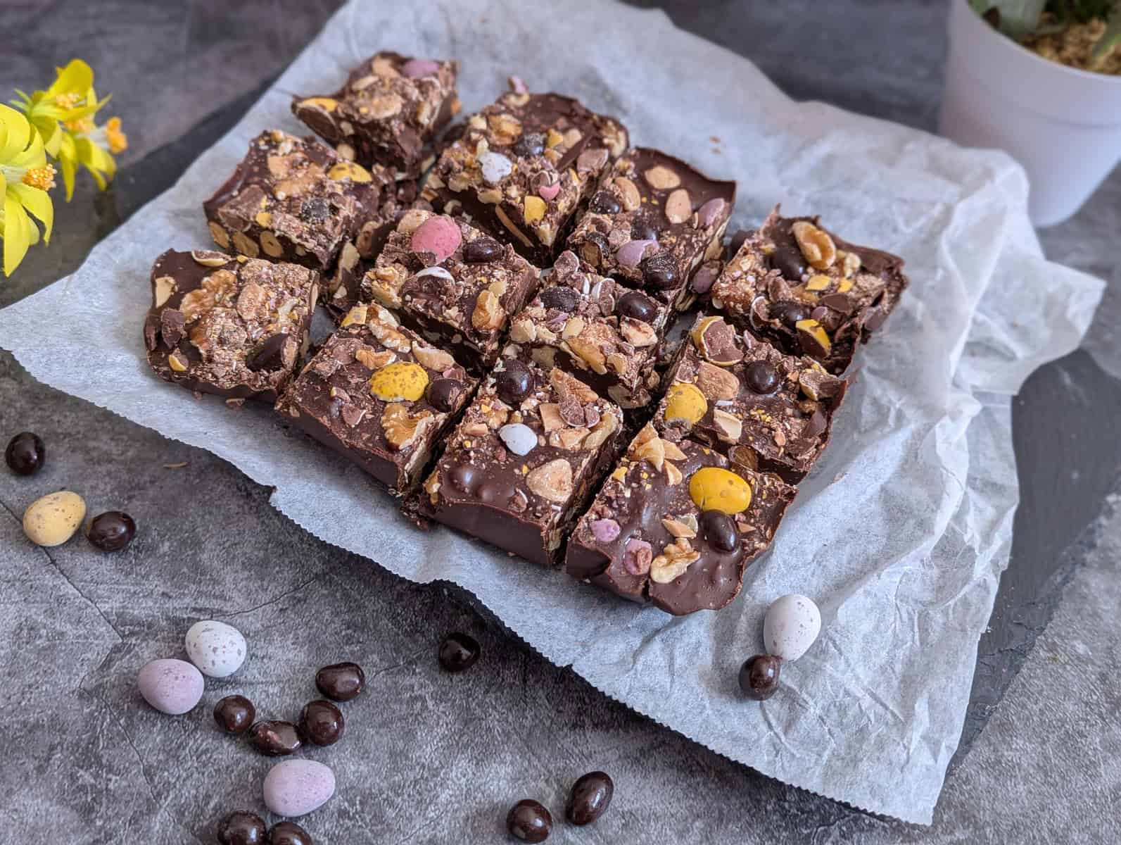 Squares of Rocky Road chocolate clusters, loaded with mini eggs, crunchy chocolate peanuts, and nuts, displayed with spring flowers and colourful Easter goodies. 