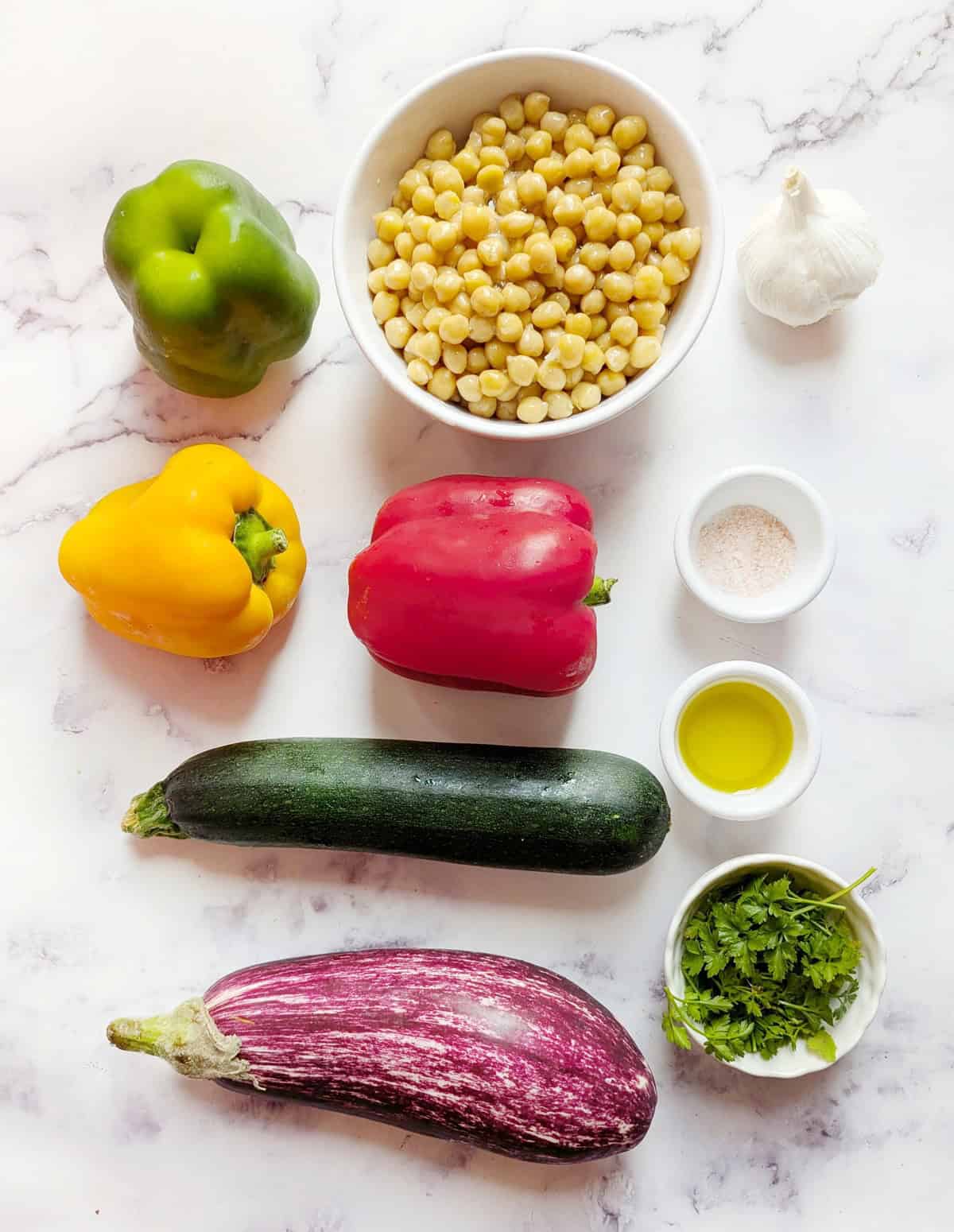 Recipe ingredients laid out on a white marble surface: red, green, and yellow peppers, cooked chickpeas, a head of garlic, fresh parsley, olive oil, eggplant, and zucchini.