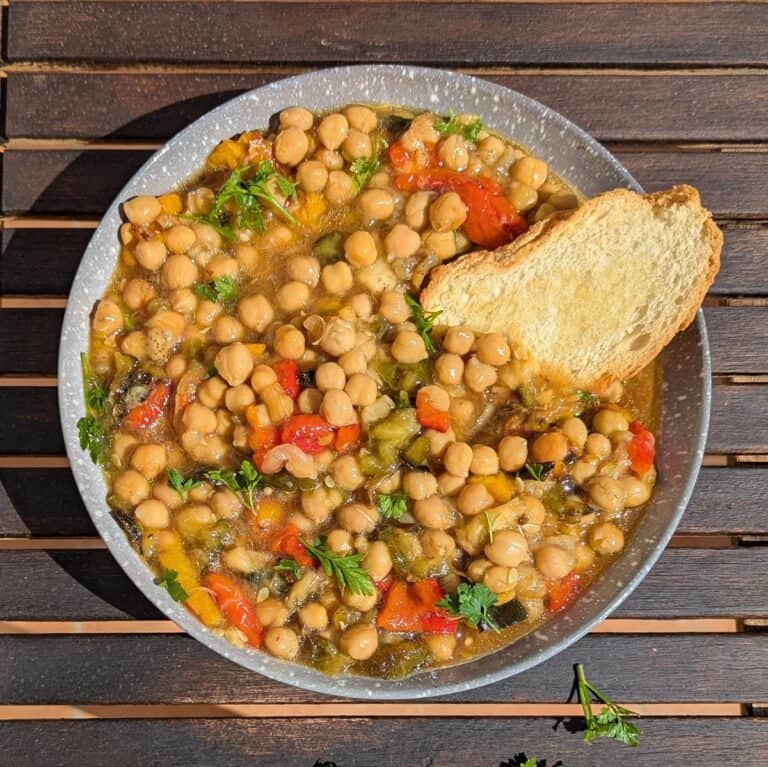 A bowl full off chickpeas with roasted vegetables and toasted bread on a wooden table with slats.