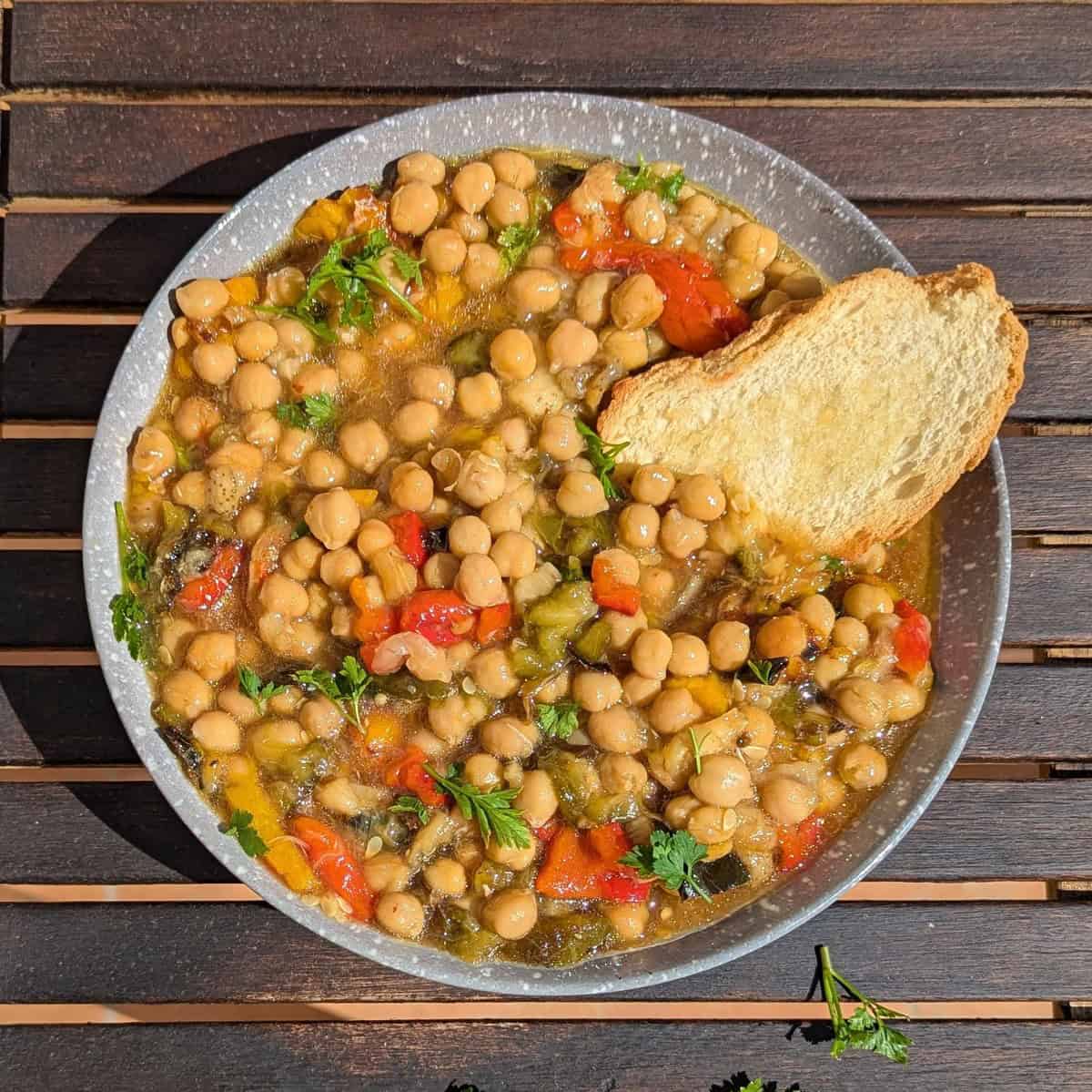 A bowl full off chickpeas with roasted vegetables and toasted bread on a wooden table with slats.