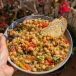 A bowl full off chickpeas with roasted vegetables and toasted bread. Faded pink flowers are in the background.