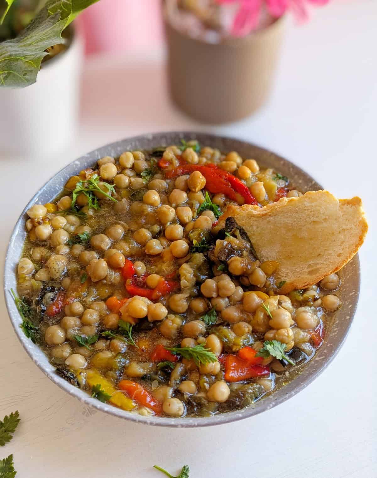 A bowl full off chickpeas with roasted vegetables and toasted bread. A faded pink plant is in the background.