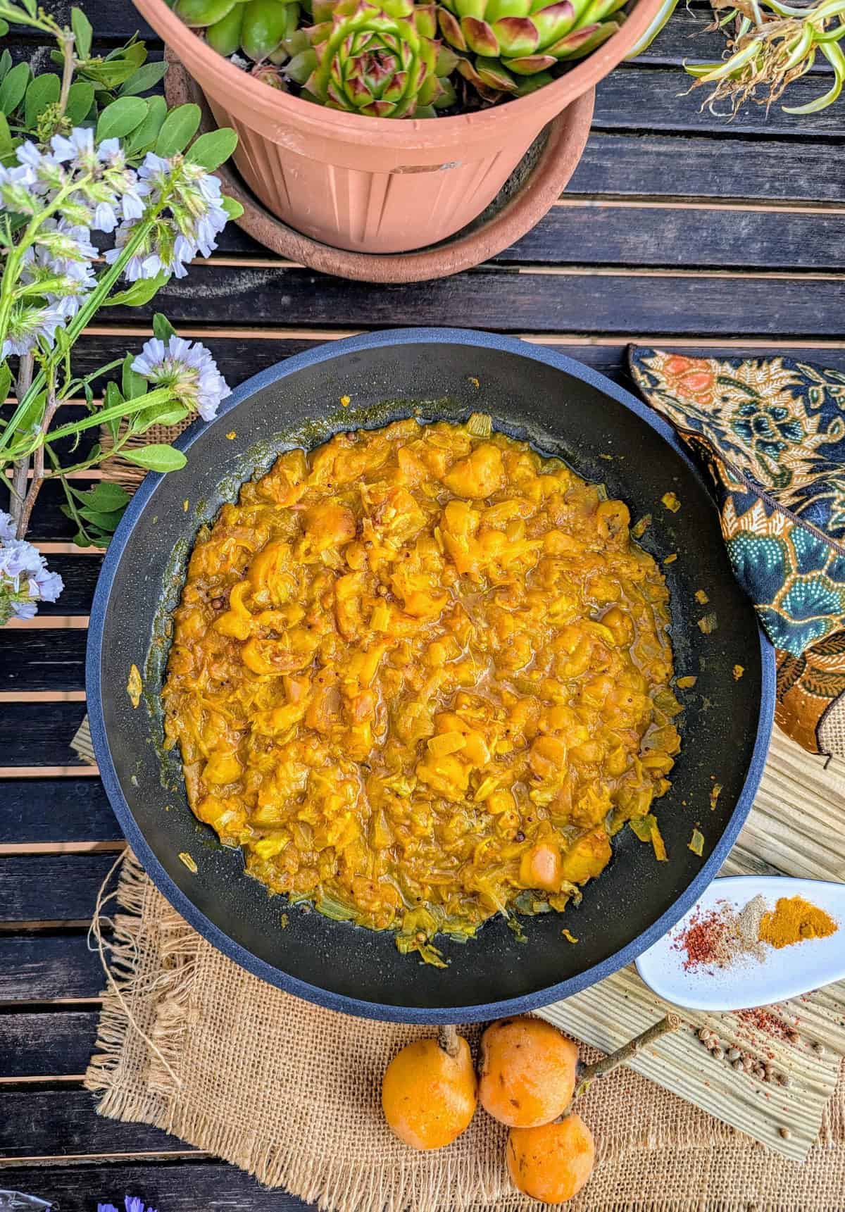 Skillet pan of freshly cooked homemade níspero (loquat) chutney with visible spices, surrounded by loquats, herbs, and a rustic setting.