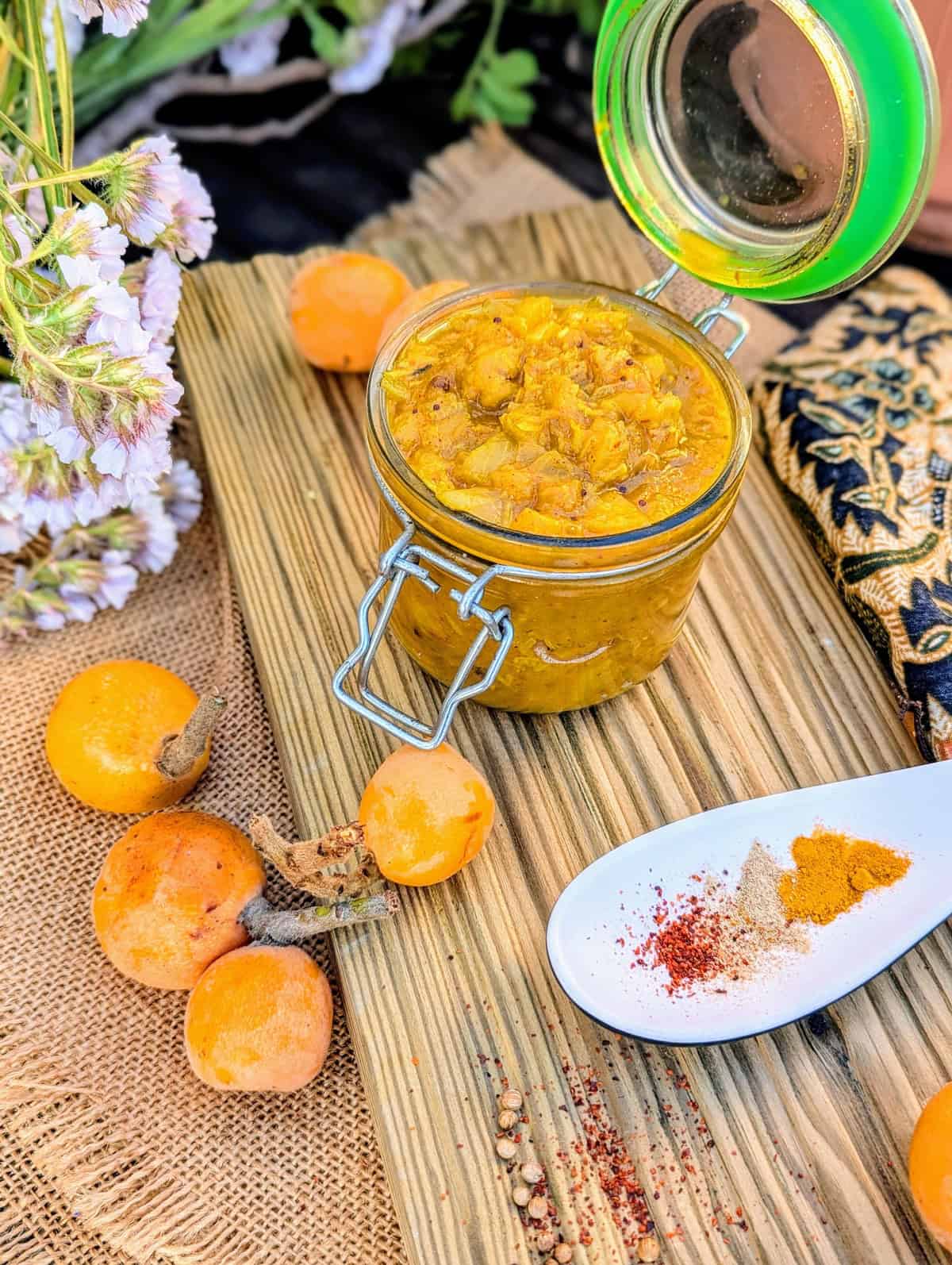 Jar of homemade níspero (loquat) chutney with fresh loquats and Indian spices on a rustic wooden board. Flowersand a pretty cloth are in the background.