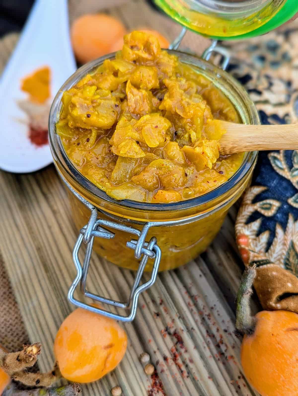 A close up Jar of homemade níspero (loquat) chutney with fresh loquats and Indian spices on a rustic wooden board.