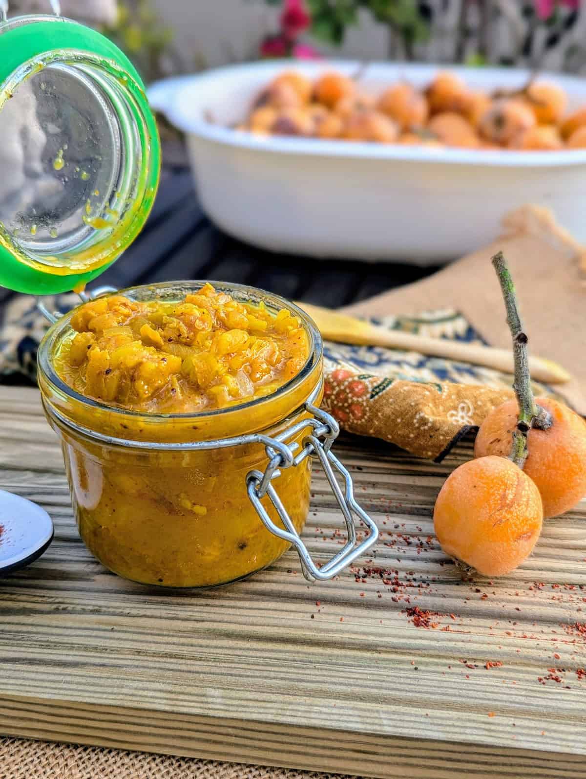 Jar of homemade níspero (loquat) chutney with fresh loquats on a rustic wooden board.