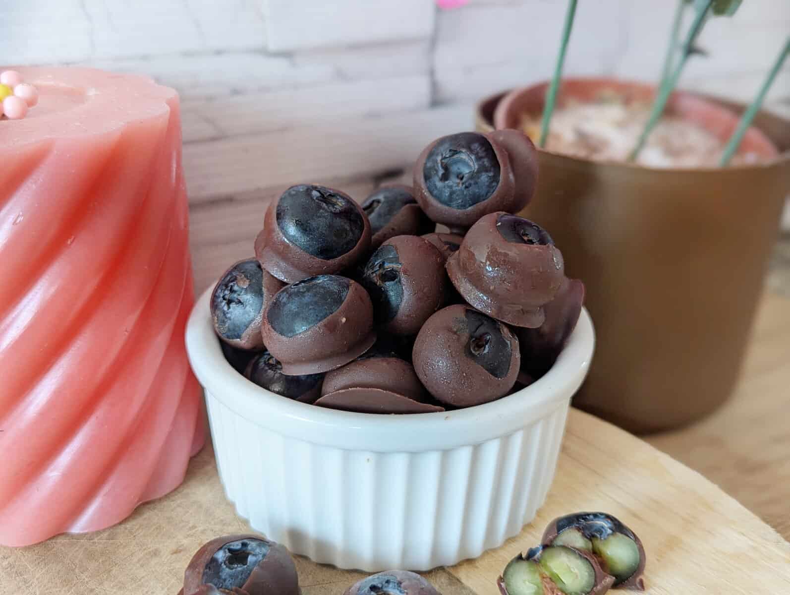 A white bowl of chocolate dipped blueberries. A pink candel and brown plant pot are in the background.