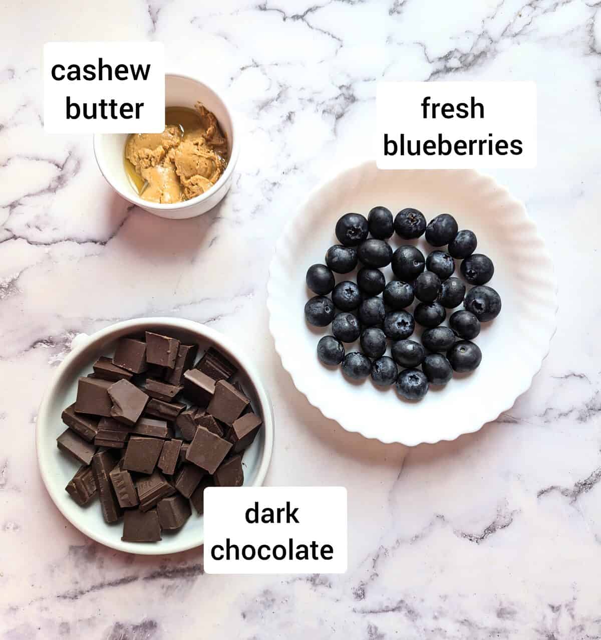 3 ingredients on a marble worktop. A bowl of chocolate squares, cashew butter and juicy blueberries.
