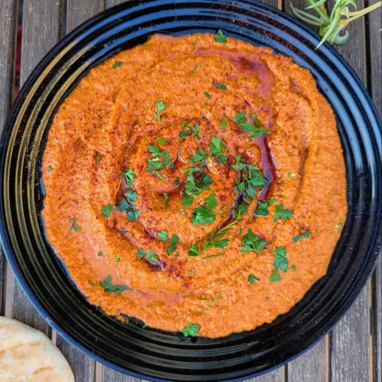 Plated is a unsweetened muhammara sprinkled with parsley, oil and paprika. A rustic table showing a pitta bread and a plant.