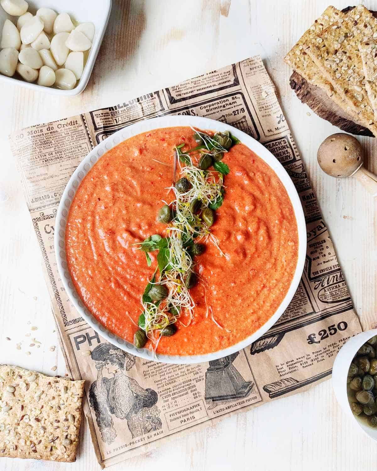A decorated bowl of red pepper hummus with crackers, capers and pickled garlic.