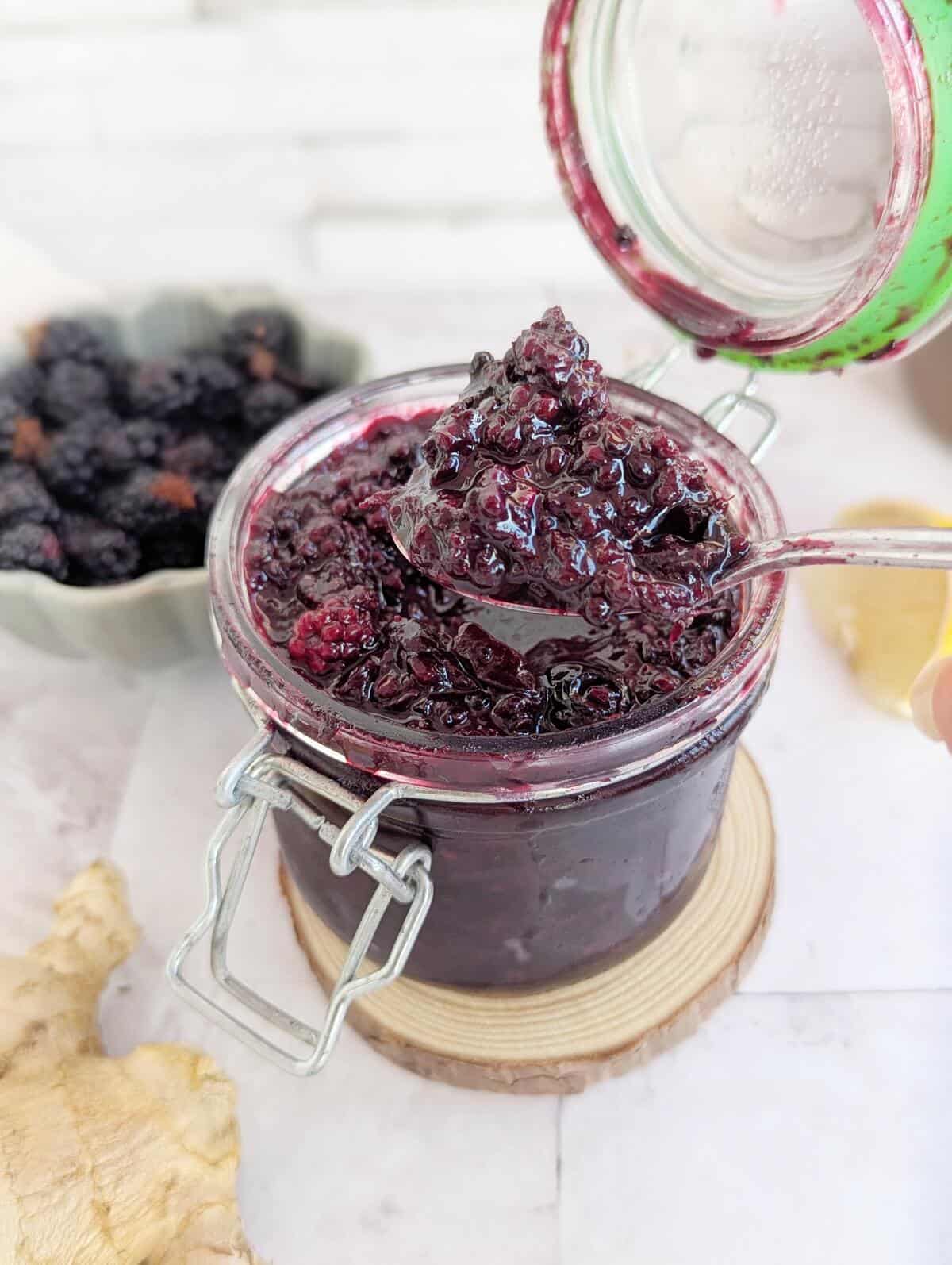 An open jar of blackberry ginger compote. Blackberries are faded in the backgroung and ginger by the side.