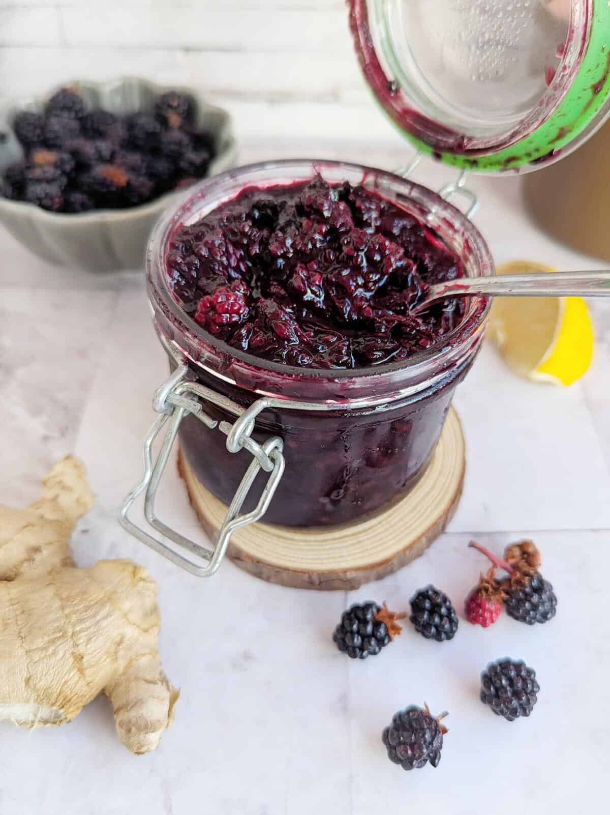 An open jar of blackberry ginger jam. Blackberries on table infront, ginger by the side and a slice of lemon behind.