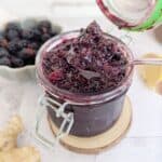 An open jar of blackberry ginger compote. A bowl of blackberries are faded in the background.