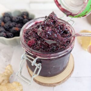 An open jar of blackberry ginger compote. A bowl of blackberries are faded in the background.
