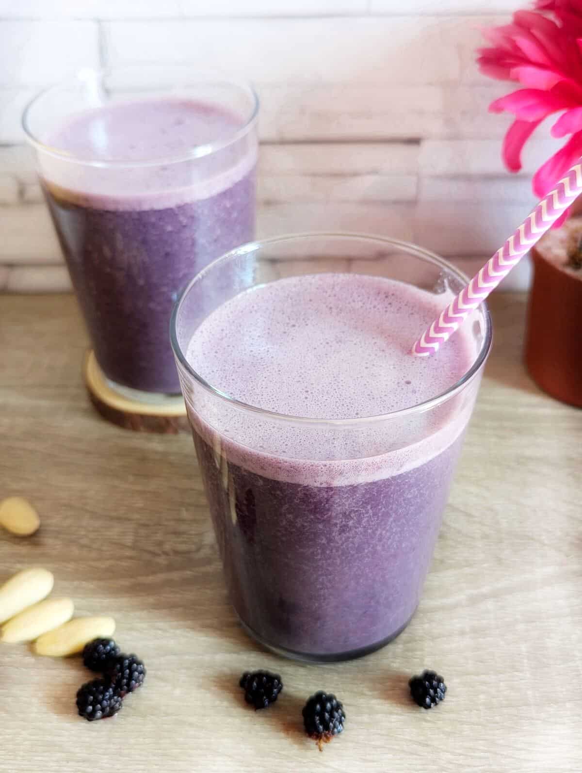 Two glasses of blackberry almond smoothie. One with a straw, and a few blackberries and almonds are lying in front.