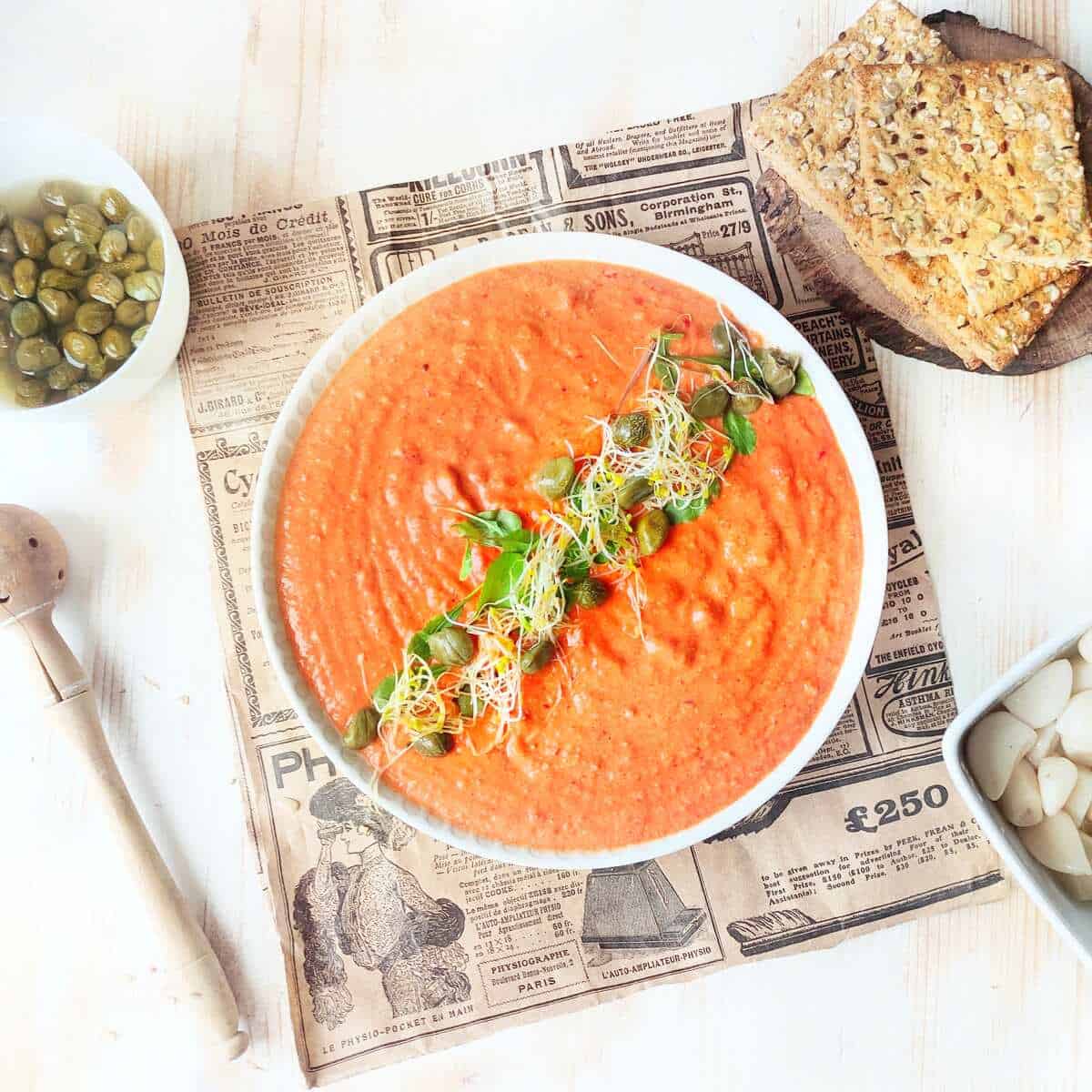 A decorated bowl of red pepper hummus with crackers, capers and pickled garlic. displayed by the side.