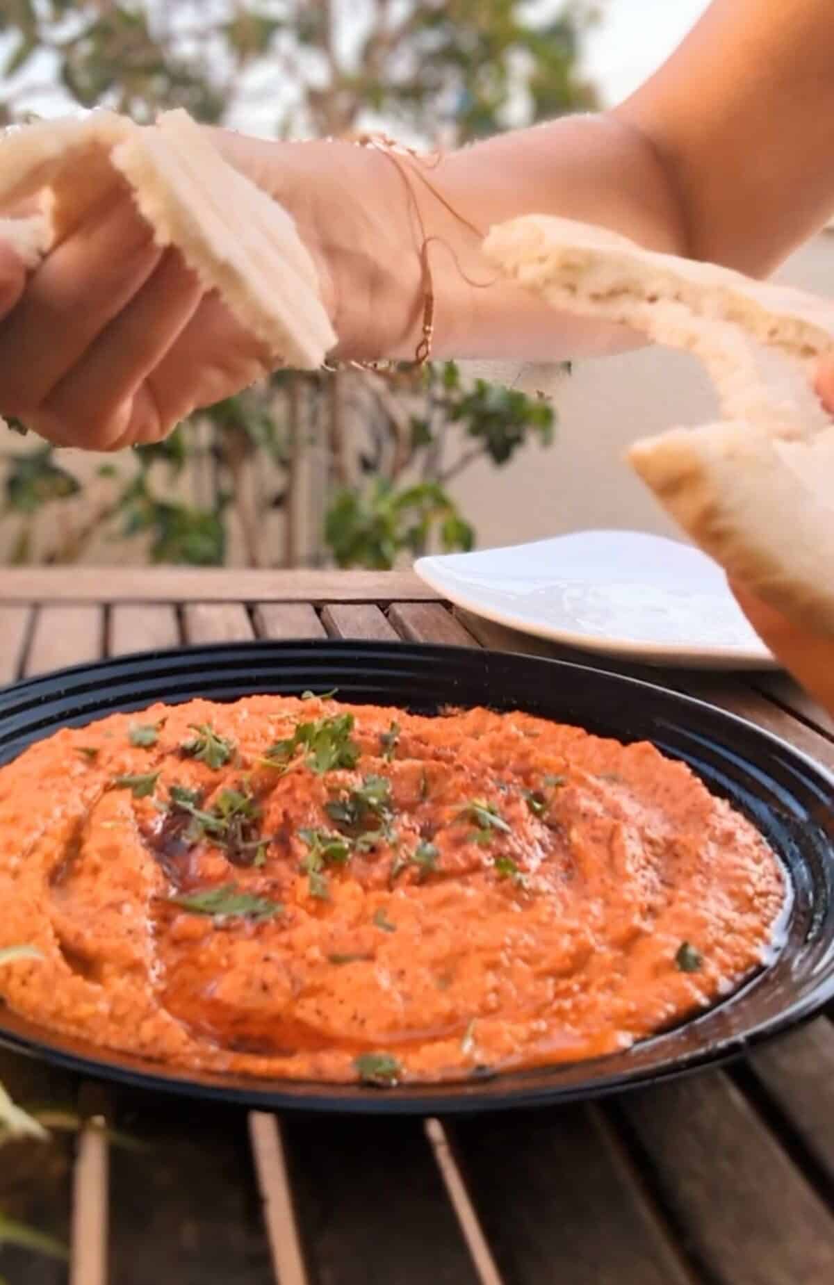 A red pepper dip sprinkled with parsley and sweet pimenon. A hand with pita bread is above the plate.
