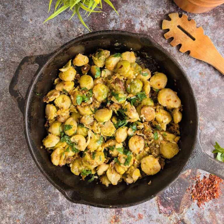 A skillet pan with sprouts and parsley. A wooden spoon lies on te right. Chili flakes are sprinkled infront.