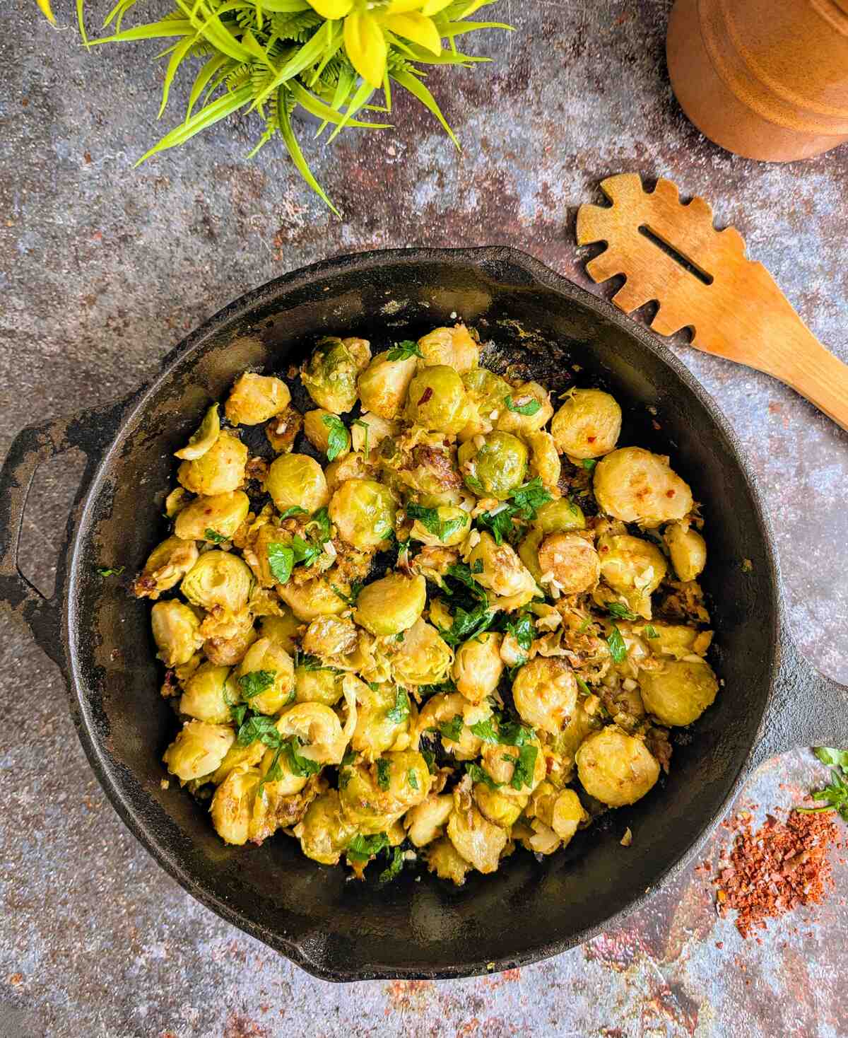 A skillet filled with brussels sprouts and herbs. A wooden spoon by the side and chili flakes.
