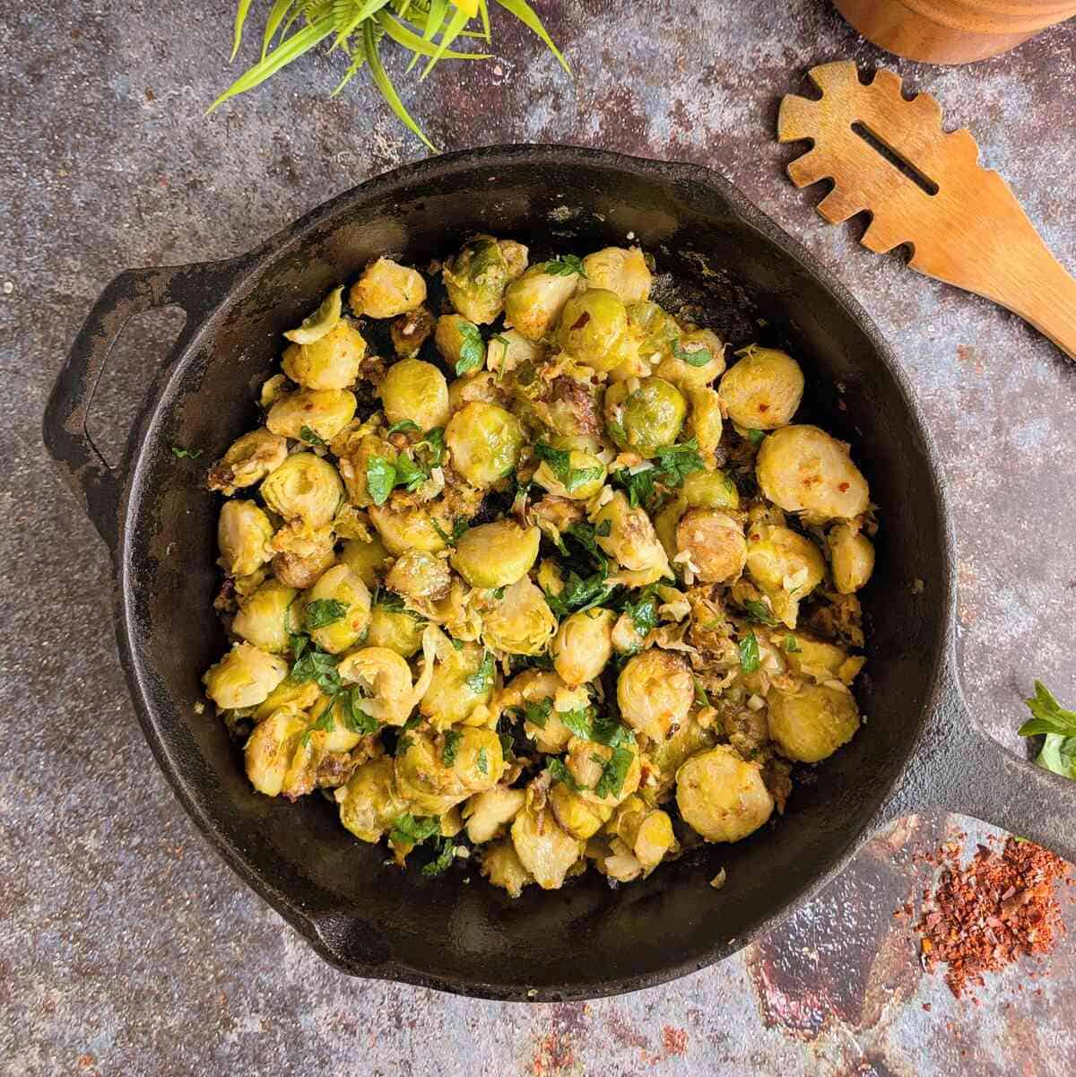 A skillet pan with sprouts and parsley. A wooden spoon lies on te right. Chili flakes are sprinkled infront.